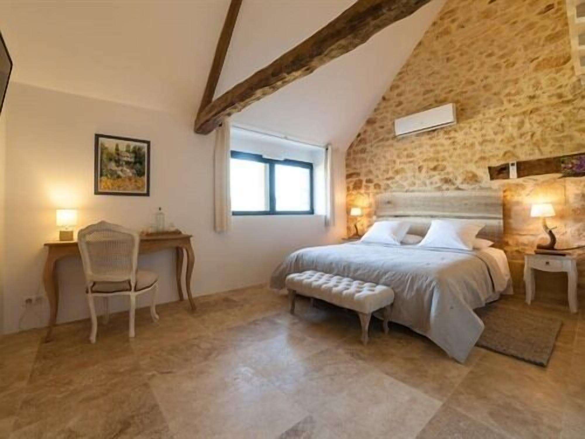 A bedroom with a stone wall, a bed with a headboard, a chair, a desk, and a window.