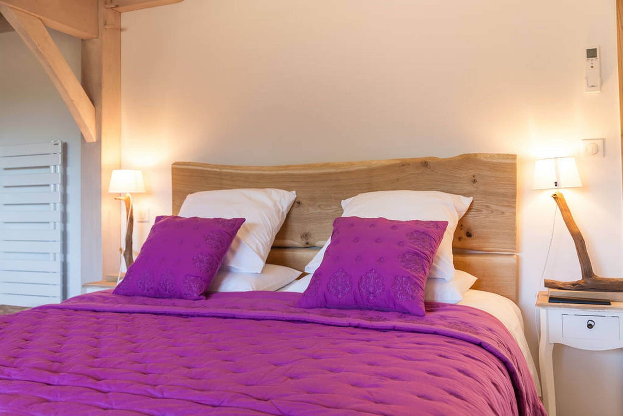 A bed with a wooden headboard, two white pillows, and two purple pillows. There are two bedside lamps on either side of the bed.