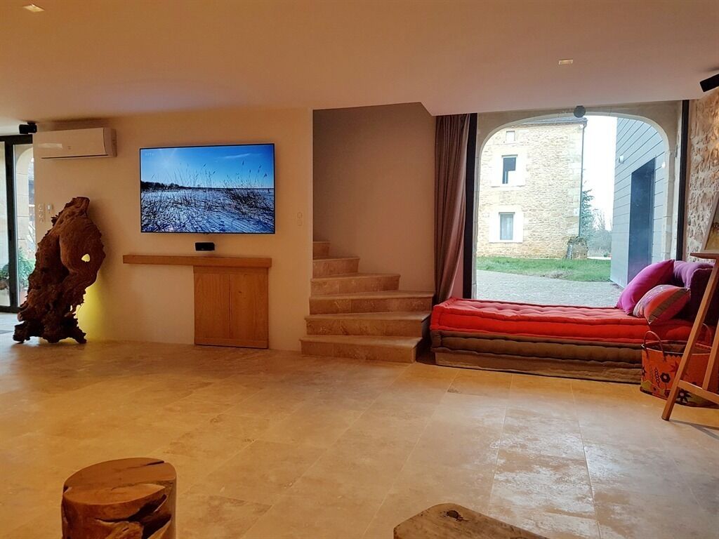 Un vaste salon doté d’un grand téléviseur à écran plat fixé au mur, d’une console en bois, d’un escalier menant à un espace extérieur et d’un coin salon confortable agrémenté d’un coussin rouge.