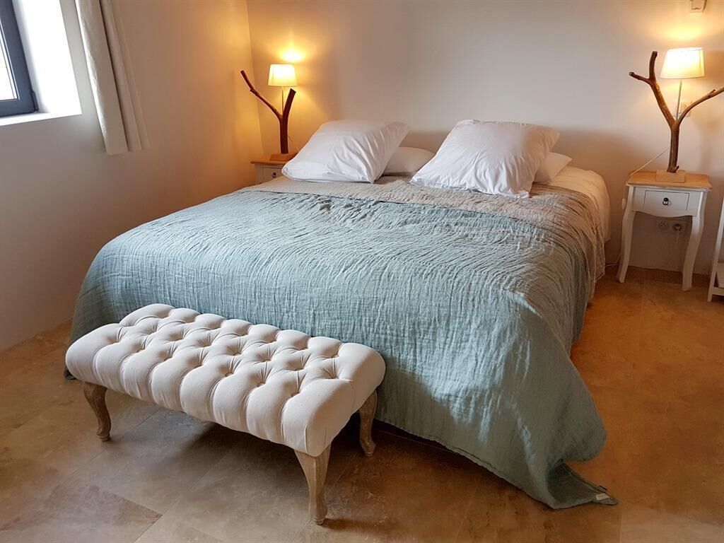 Un dormitorio con una cama grande, dos mesitas de noche, un banco y una ventana con cortinas.