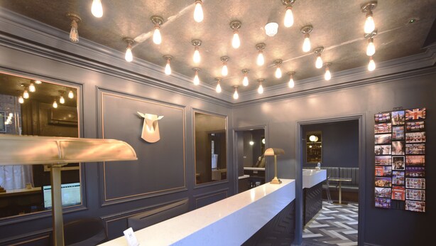 A hotel reception area with a long counter, multiple light fixtures, a mirror, and a wall with a collection of photographs. - House of Toby (London)