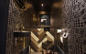 Un baño estrecho, tenuemente iluminado, con un papel tapiz a cuadros de galgo, un grifo dorado, un lavamanos negro y un inodoro con tapa negra.