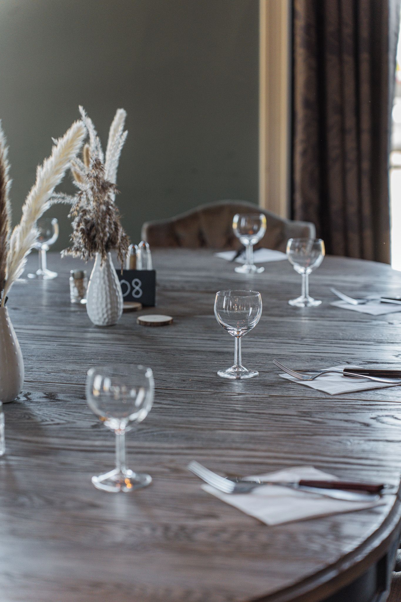 A wooden dining table with several wine glasses and a vase with dried plants.