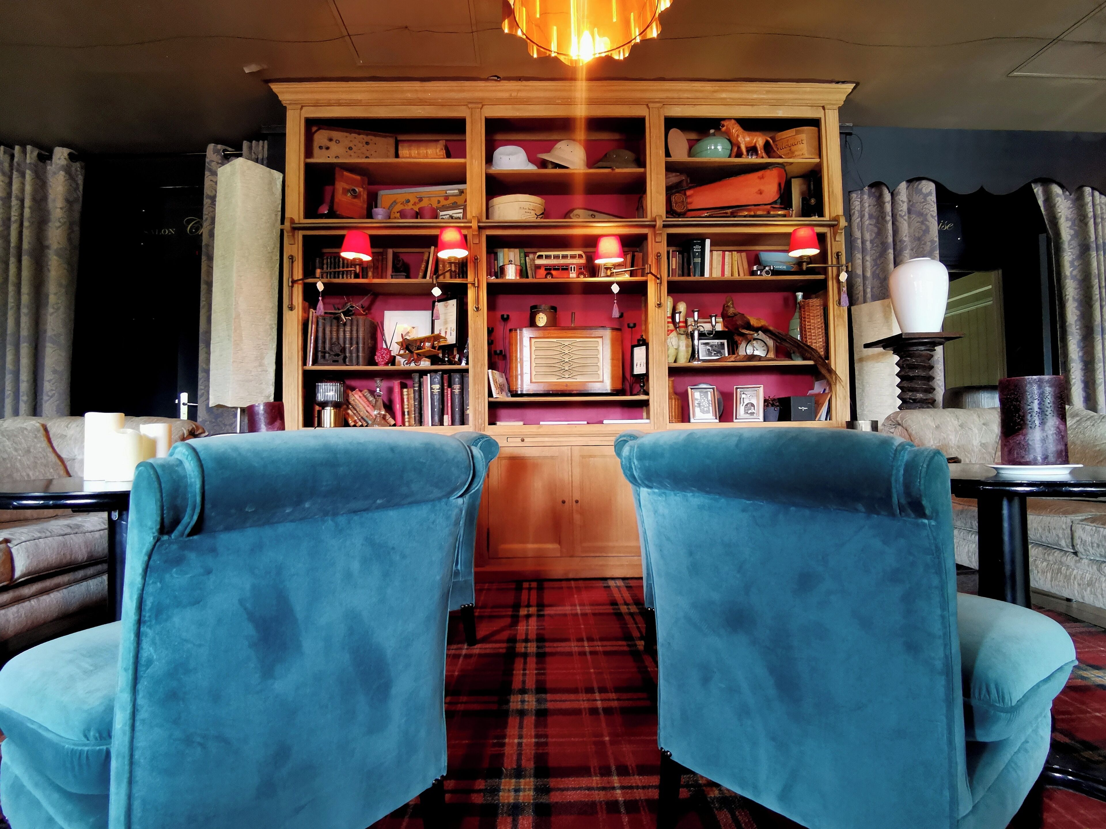Une pièce avec deux fauteuils bleus, une bibliothèque en bois remplie de livres et d’objets décoratifs, et un tapis à carreaux rouges.