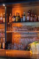 Zona de bar con estantes llenos de botellas de licores y numerosos vasos de vino.
