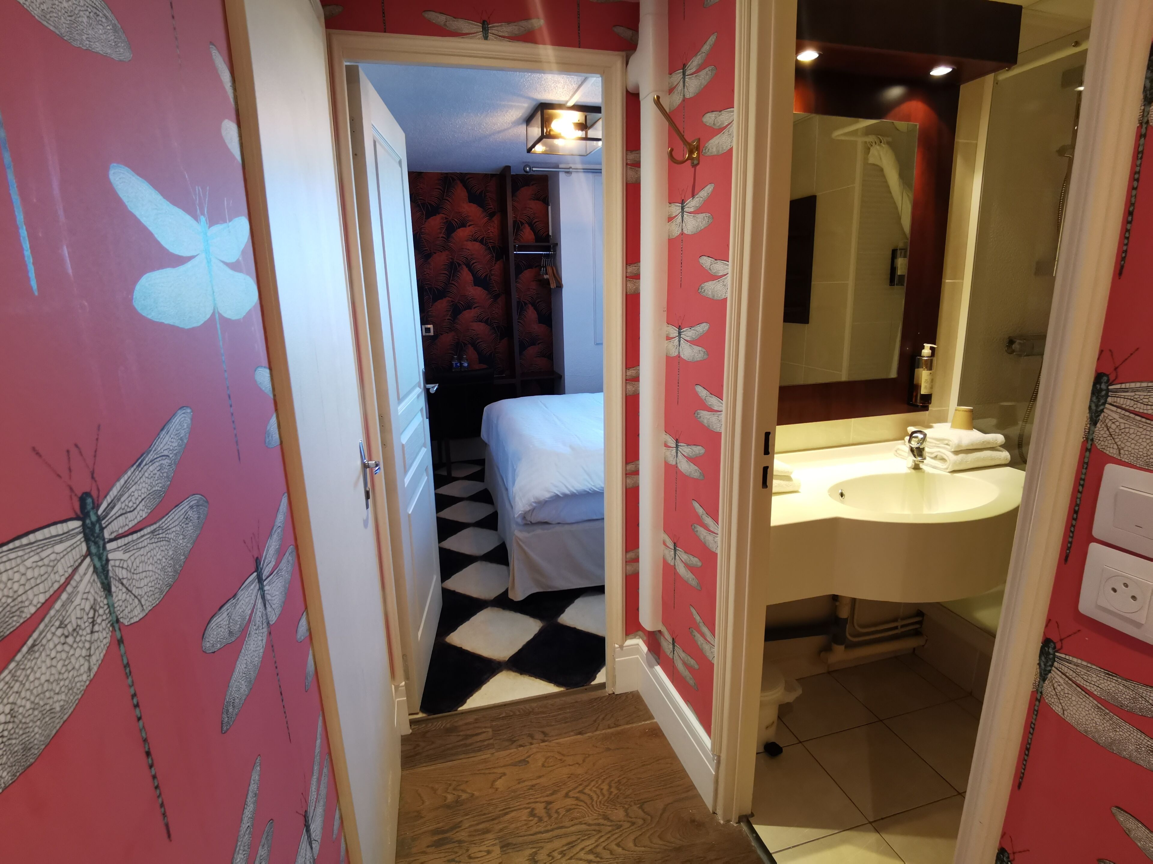 A hotel room with a checkered floor, a white bed, a bathroom with a sink and mirror, and a door leading to another room.