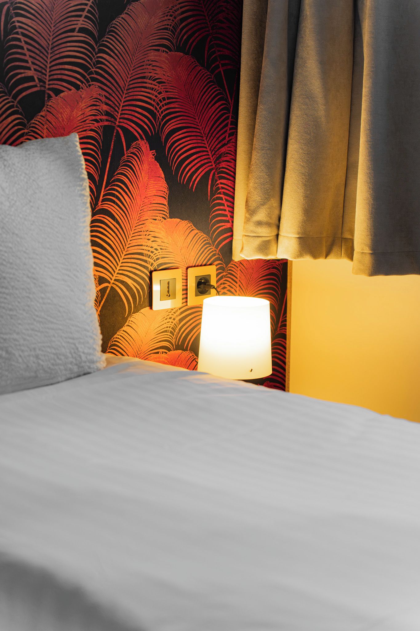 Habitación de hotel con cama, mesita de noche con lámpara y pared decorada con un estampado de hojas tropicales.