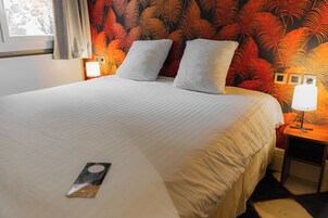 A hotel room with a large bed, two pillows, a nightstand with a lamp, and a wall with a tropical leaf pattern.