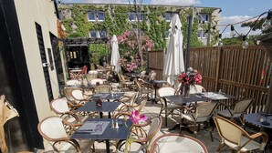 An outdoor café with round tables and wicker chairs, surrounded by a wooden fence and greenery. - Hôtel Mercator (Vendome)