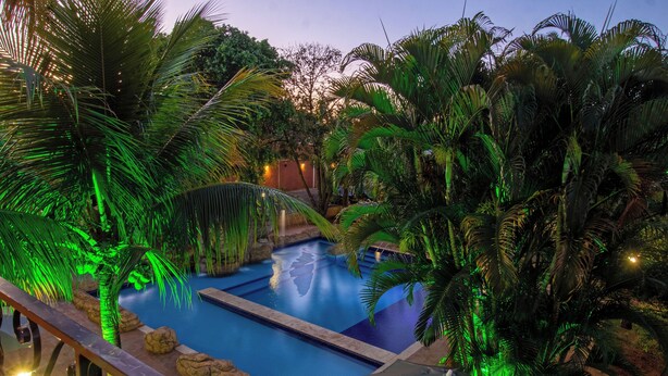 A swimming pool surrounded by tropical plants and rocks, with a view of the sky. - Pousada Faruk (Olímpia)