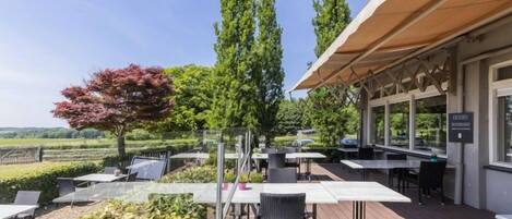 An outdoor patio area with tables and chairs, surrounded by greenery and tall trees.