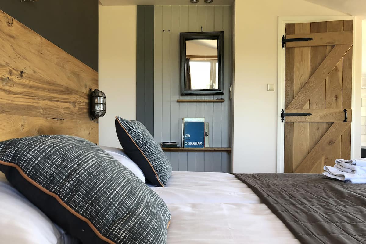 Un dormitorio con cabecera de madera, cama grande con almohadas estampadas, mesita de noche con un libro y una puerta de madera.
