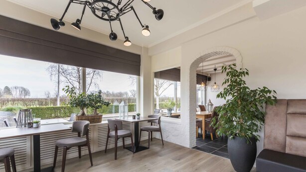 A modern dining area with a long table and chairs, large windows, and a potted plant. - Hotel de Plank (Noorbeek)