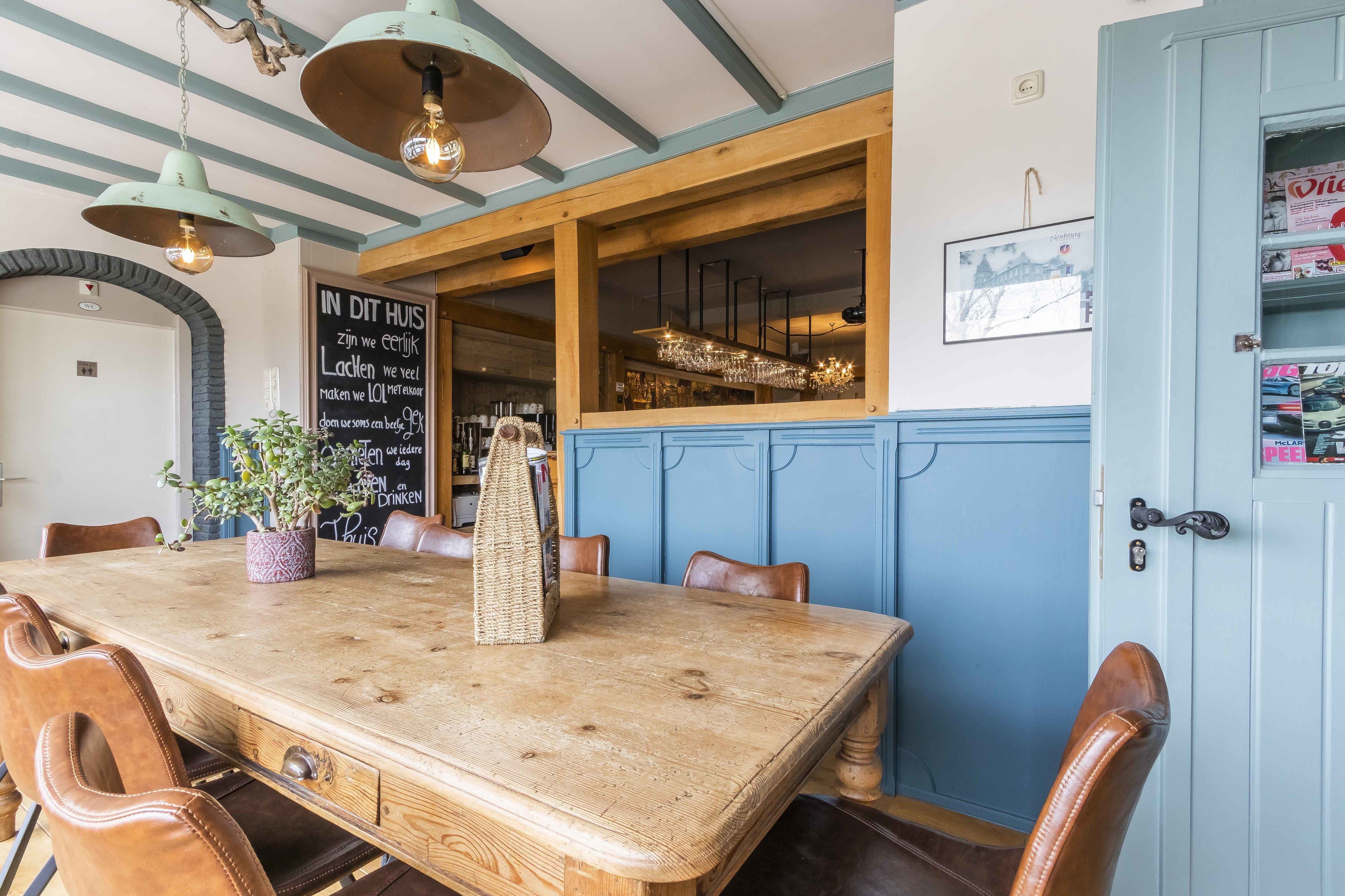 Una mesa de comedor de madera con sillas, una planta en maceta y lámparas colgantes en una habitación con paredes azules y vigas de madera.