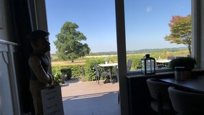 A view from inside a room with a large window overlooking a patio area with a table and chairs, a sign with text, and a statue. - Hotel de Plank (Noorbeek)