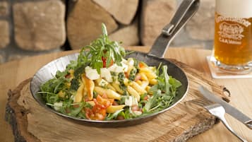 A plate of pasta with vegetables and cheese, a fork, and a glass of beer on a wooden table.
