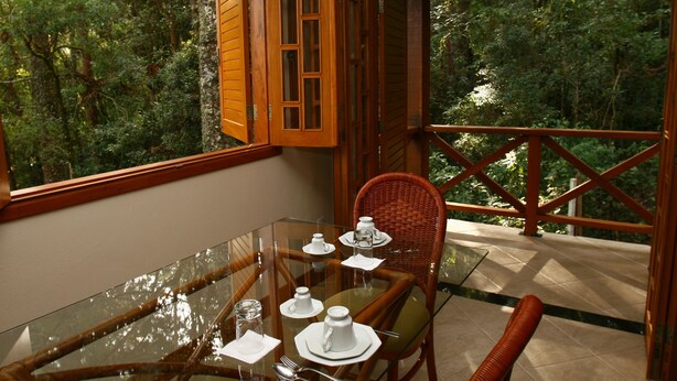 A dining area with a glass table, wicker chairs, and a view of a forest through open wooden shutters. - Chalé da Floresta (Camanducaia)