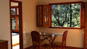 A dining area with a glass table and wicker chairs, a wooden cabinet with glass doors, and a window with a view of trees. - Chalé da Floresta (Camanducaia)