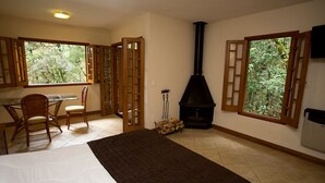A bedroom with a bed, a desk, chairs, a fireplace, and large windows with a view of greenery. - Chalé da Floresta (Camanducaia)