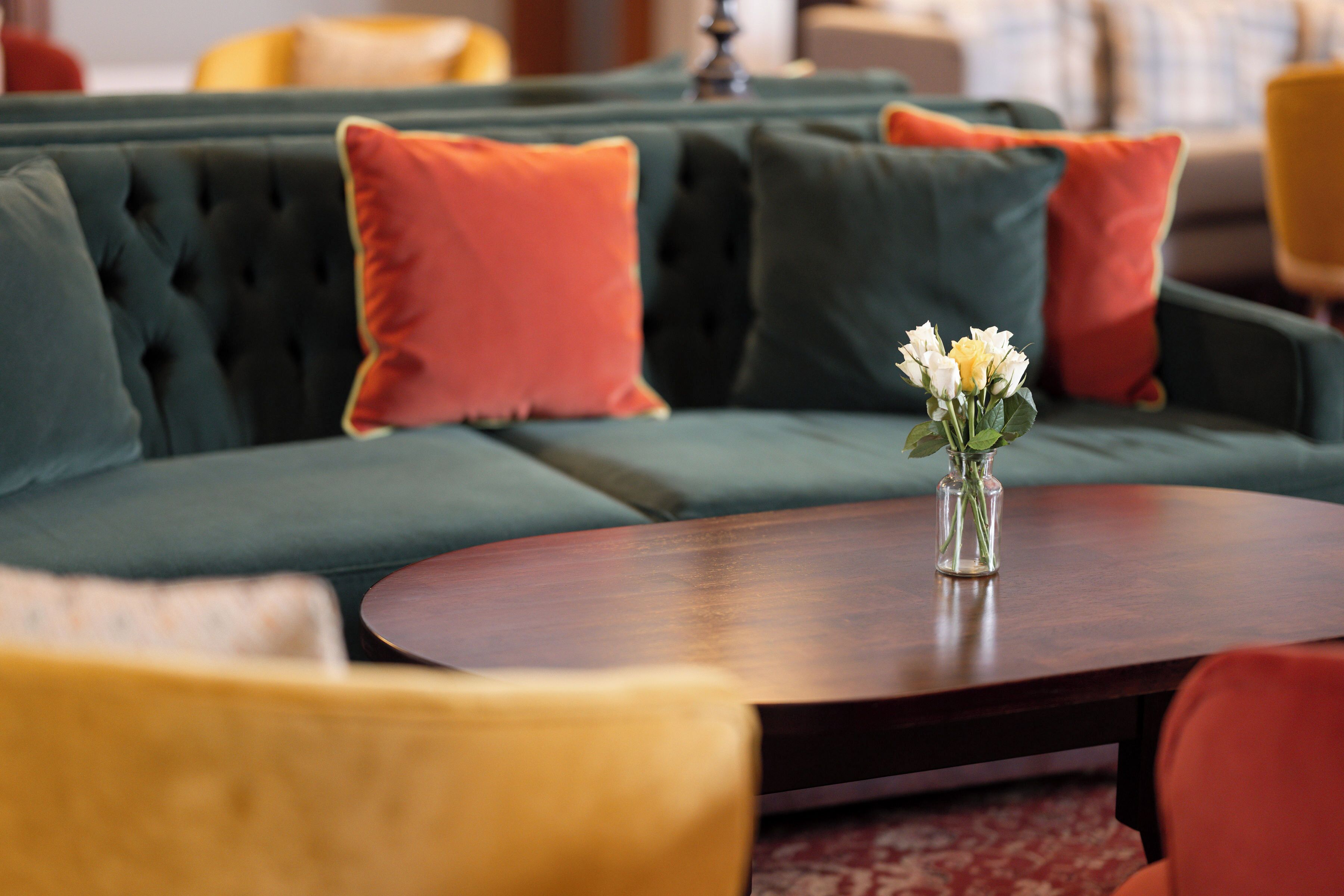 Un canapé vert recouvert de tissu, agrémenté de coussins orange, une table basse en bois et un vase avec des fleurs blanches.