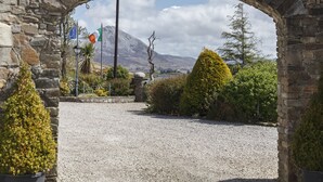 A stone archway leading to a gravel path with a mountain in the background. Flags of the EU and Ireland are visible. - An Chúirt Hotel (Gweedore)