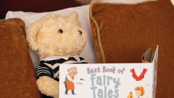 Un oso de peluche con un libro titulado "Best Book of Fairy Tales" sobre una cama con almohadillas marrones y una pared con diseño de fondo.