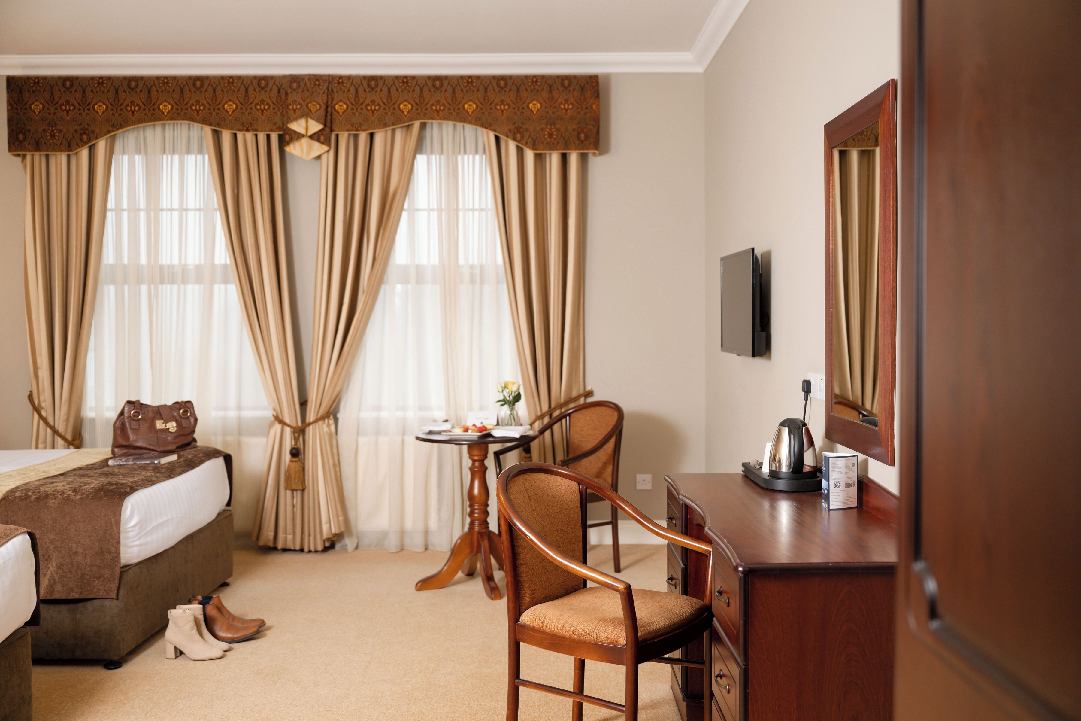 Habitación de hotel con cama, escritorio, silla, mesita con un florero y una tetera, y cortinas.