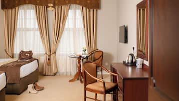 Habitación de hotel con cama, escritorio, silla, mesita con un florero y una tetera, y cortinas.