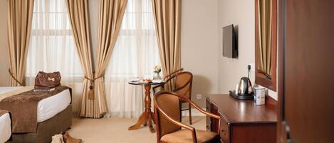 A hotel room with a bed, a desk, a chair, a small table with a vase and a kettle, and curtains.