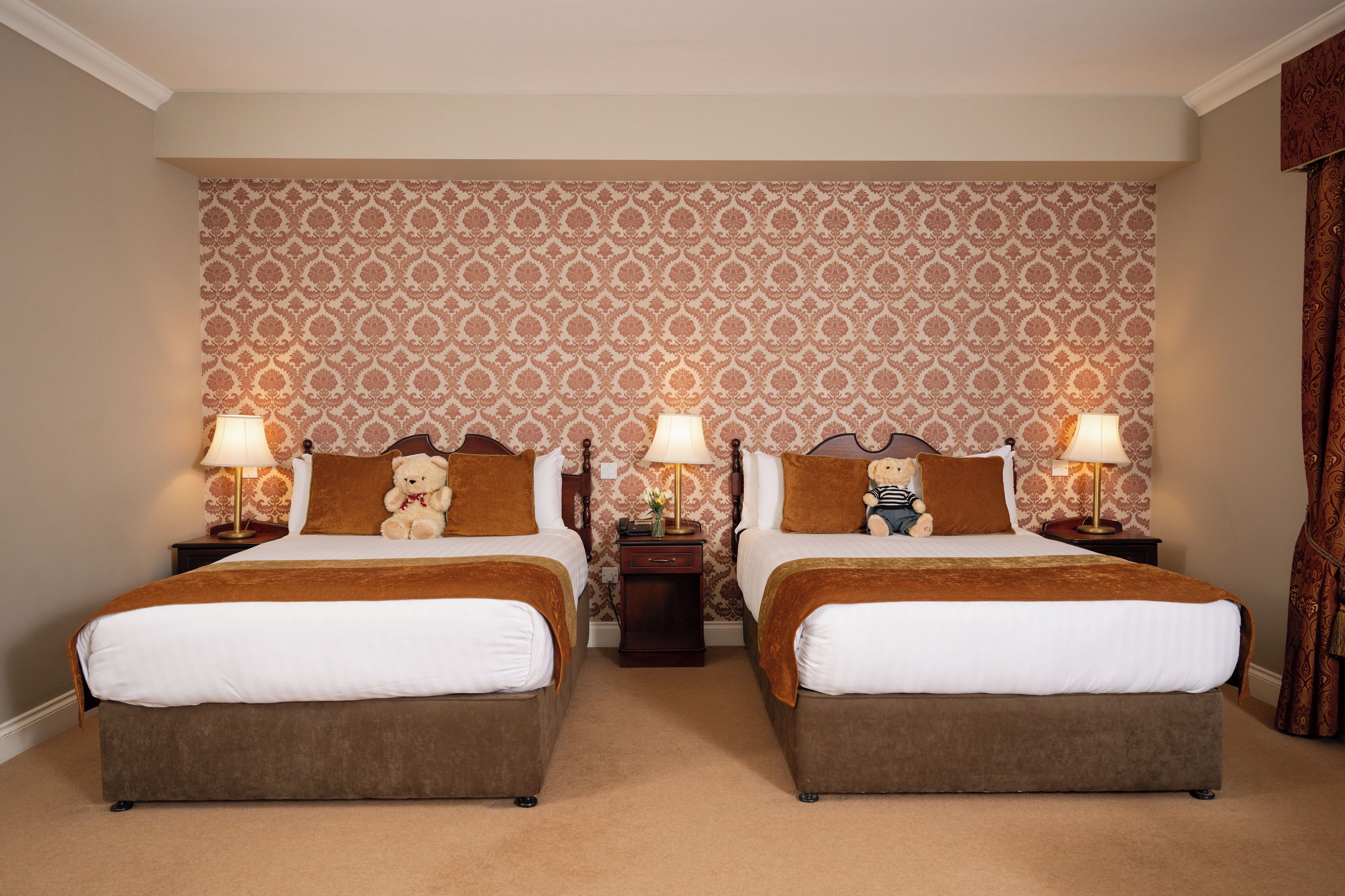 A hotel room with two beds, each with a brown headboard, white bedding, and a teddy bear on the pillows. There are two bedside tables with lamps and a small table between the beds. The room has patterned wallpaper and a carpeted floor.