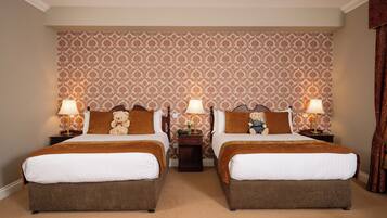 A hotel room with two beds, each with a brown headboard, white bedding, and a teddy bear on the pillows. There are two bedside tables with lamps and a small table between the beds. The room has patterned wallpaper and a carpeted floor.