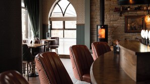 A restaurant interior with a long wooden bar, leather booth seating, a large arched window, and a stone wall with a mounted TV. - An Chúirt Hotel (Gweedore)