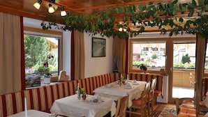 A dining area with tables set for two, a bench along the wall, and a view of the outdoors through large windows.