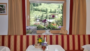 A hotel room with a large window offering a view of a green hillside and buildings. There is a framed picture on the wall, a small table with a candle, a vase with flowers, and a heart-shaped decoration on the windowsill.