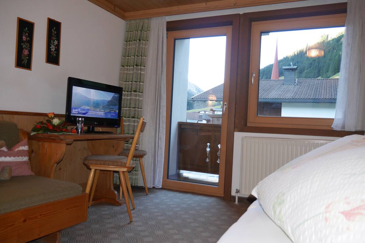 Habitación de hotel con cama, un escritorio de madera con televisor, una silla y vista a la montaña por la ventana.