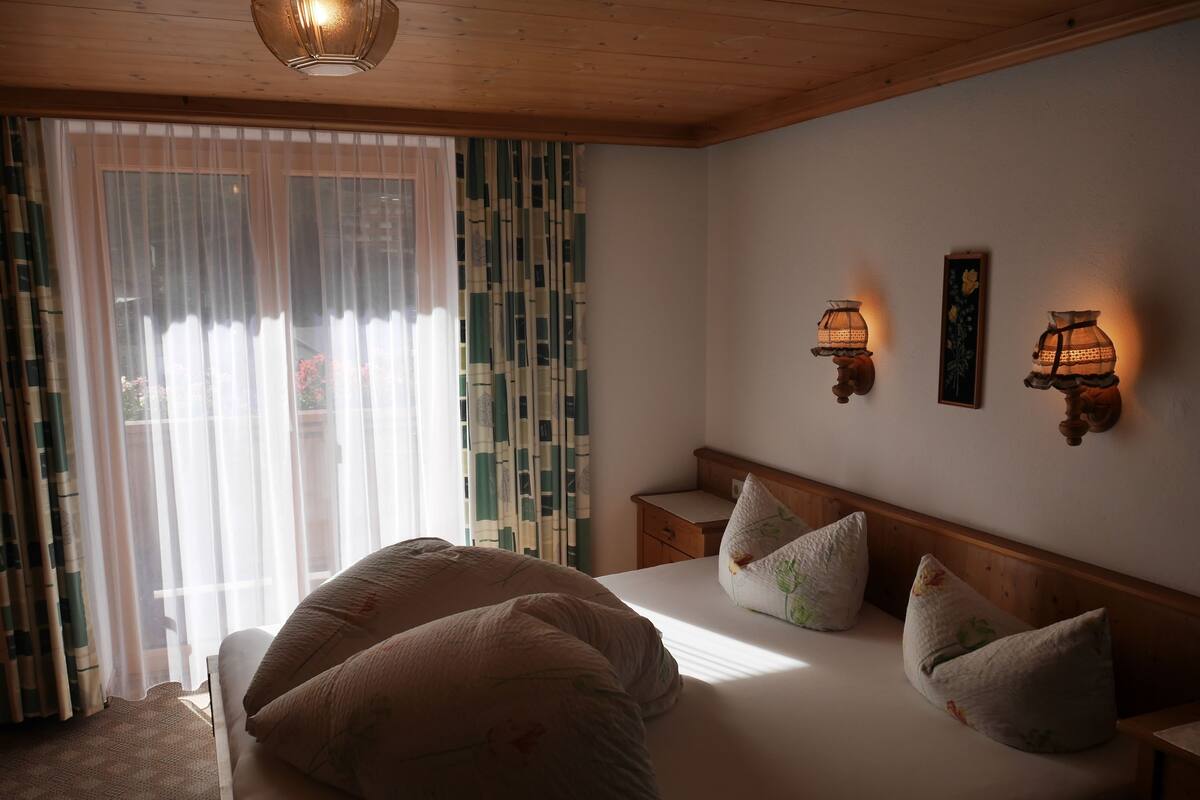 Un dormitorio con cama, almohadas, una mesita de noche, una lámpara de pared y una ventana con cortinas.