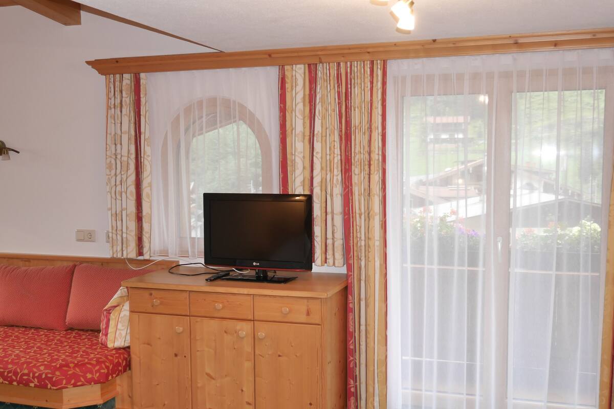 Una habitación con un armario de madera, un televisor encima, un sofá rojo y una ventana con cortinas.