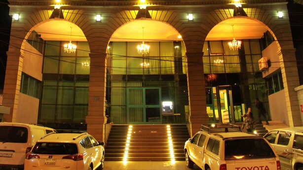 Een hotel met een opvallend bord waarop 'ZMAMA HOTEL' staat. Er zijn meerdere verdiepingen met balkons en de ingang heeft drie grote bogen met verlichting. Verschillende auto's staan ervoor geparkeerd, waaronder een Toyota.