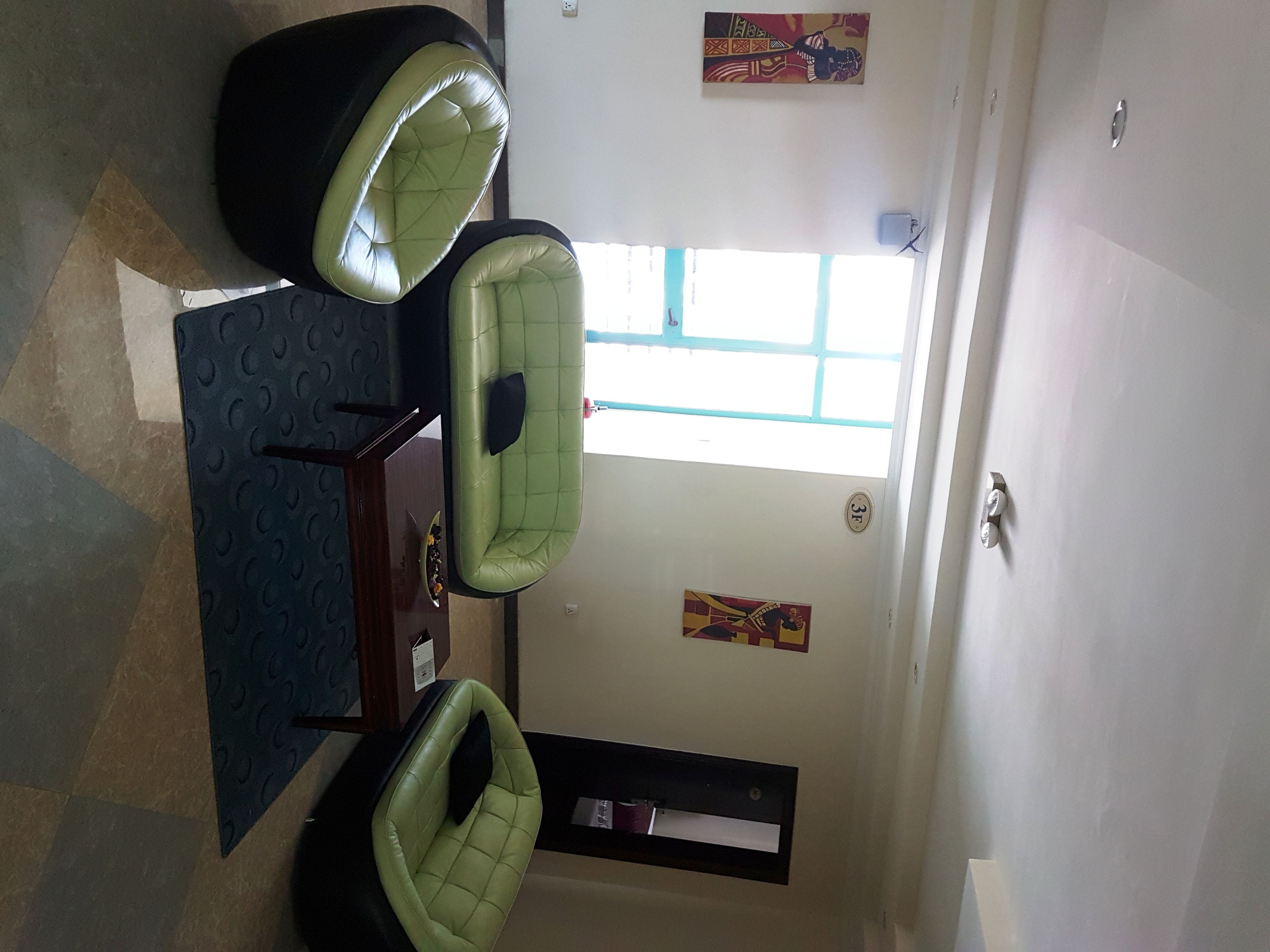 Een kamer met een eettafel en vier groene stoelen, een klein raam en een deur met een nummerbord.