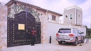 A gated entrance with a security guard, a parked SUV, and a building with a white facade and a balcony. - GGC Apartments (Lagos)