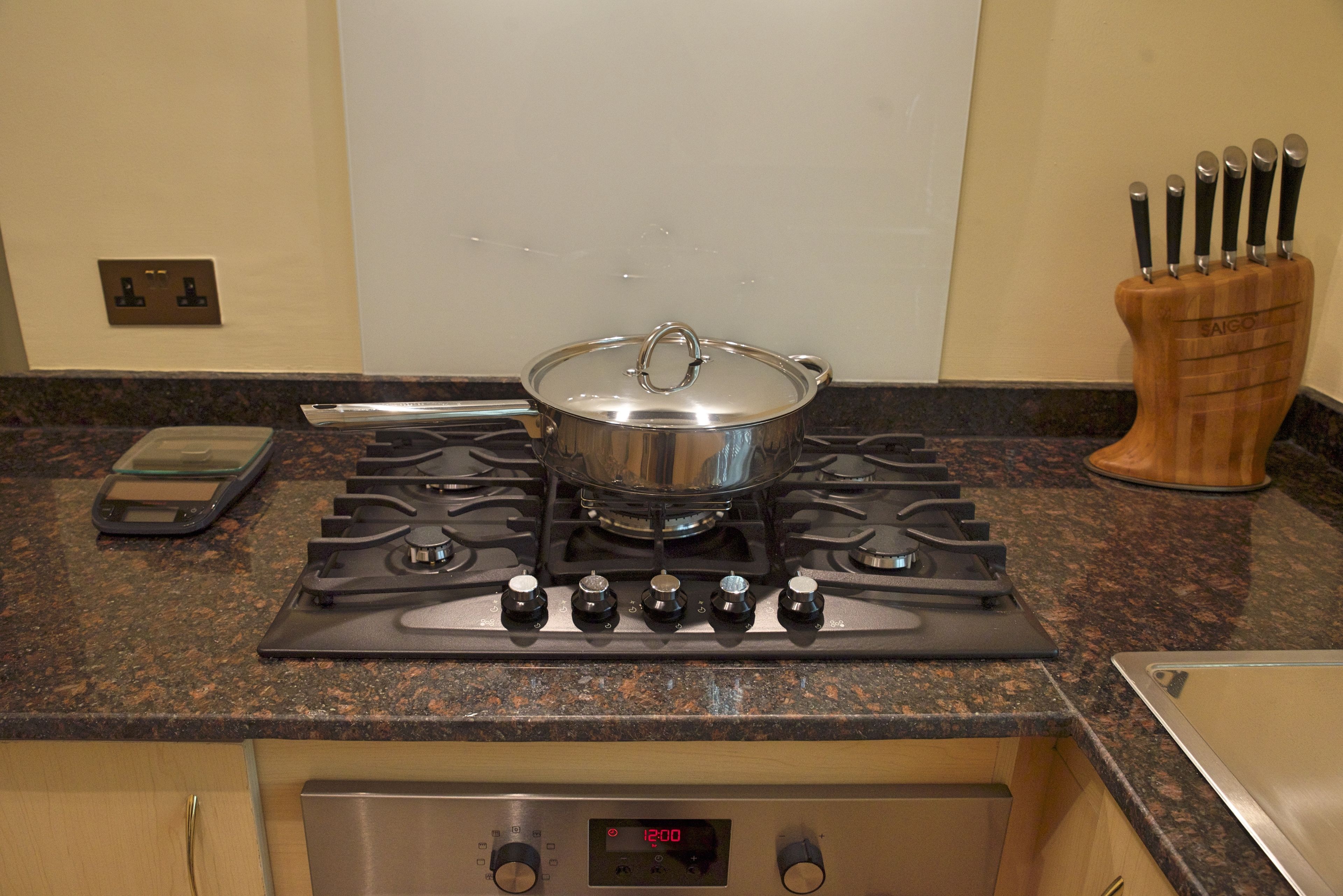A kitchen with a stove, a pot on the burner, a knife block, and a digital clock displaying 12:00.