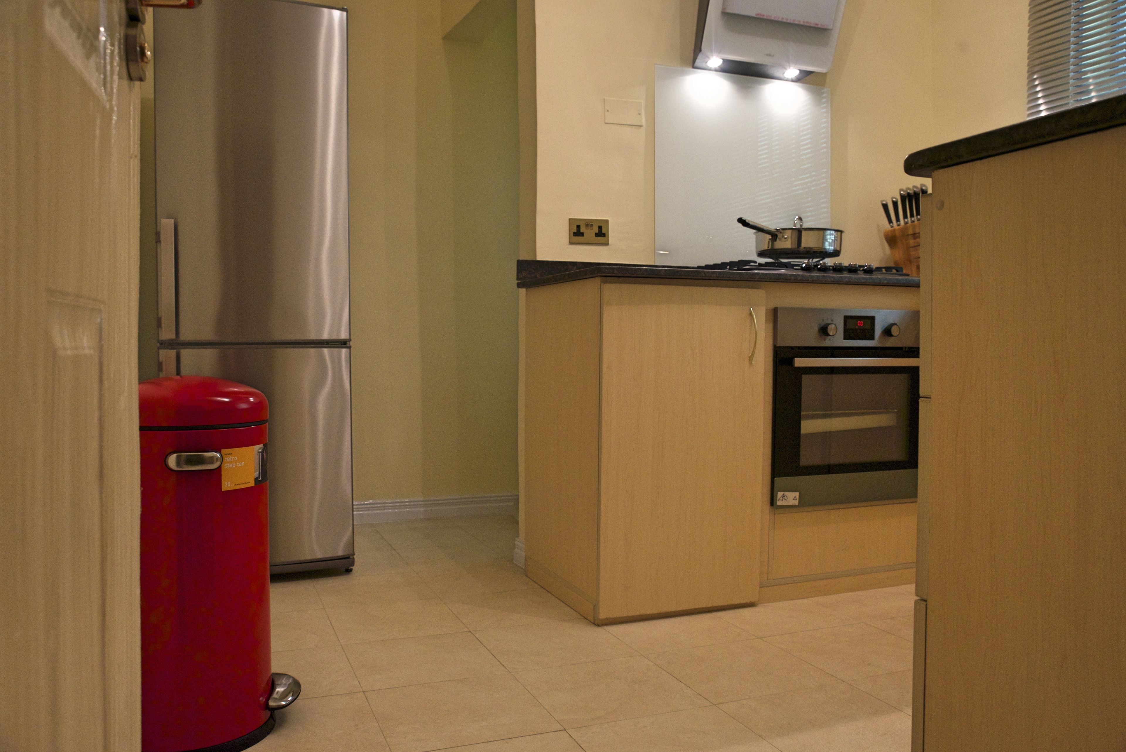 A compact kitchen with a stainless steel refrigerator, a red trash bin, a stove, and an oven.