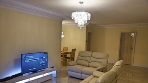 A modern living room with a flat-screen TV, a beige sofa, a dining area with a table and chairs, and a chandelier. - GGC Apartments (Lagos)