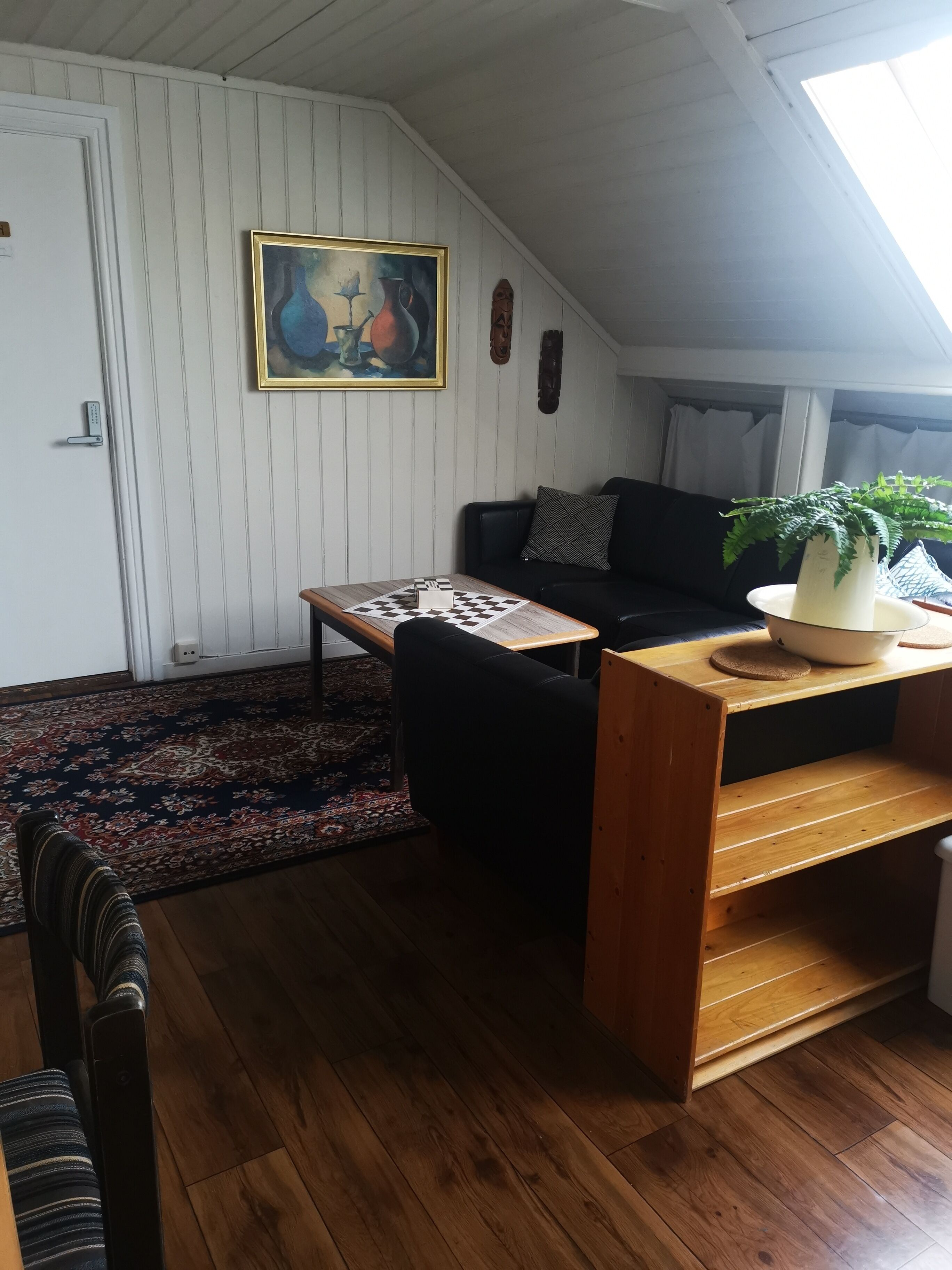 A cozy living room with a wooden floor, a black sofa, a small wooden table, a painting on the wall, and a window with curtains.