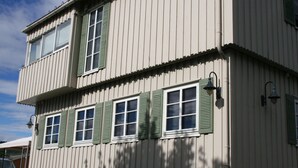 Un edificio de dos pisos con fachada sim étrica, ventanas uniformemente distribuidas con contraventanas verdes y una entrada central con puerta y un pequeño alero.