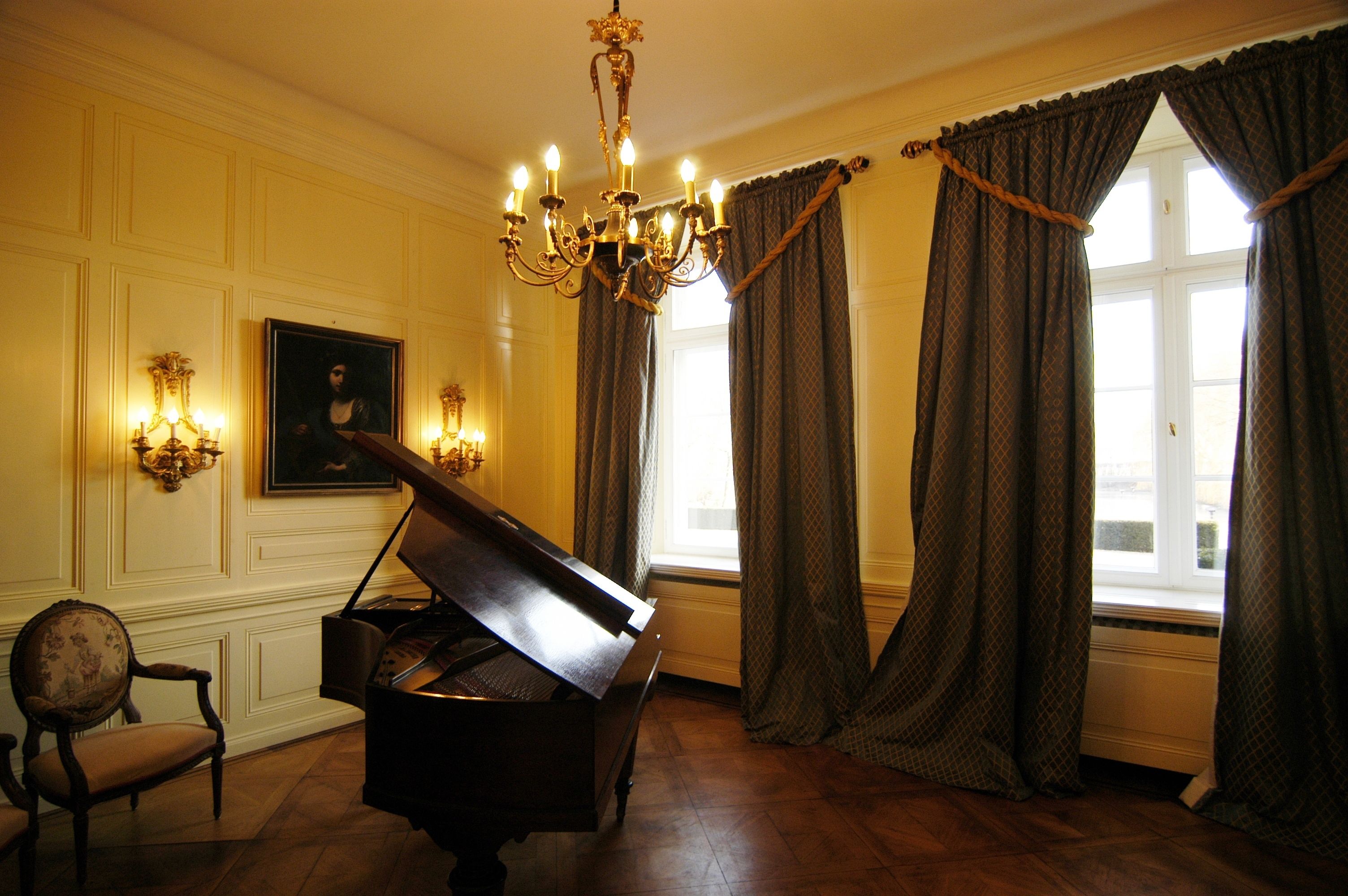 Un piano de cola en una habitación con una araña, un retrato enmarcado y una ventana con cortinas.