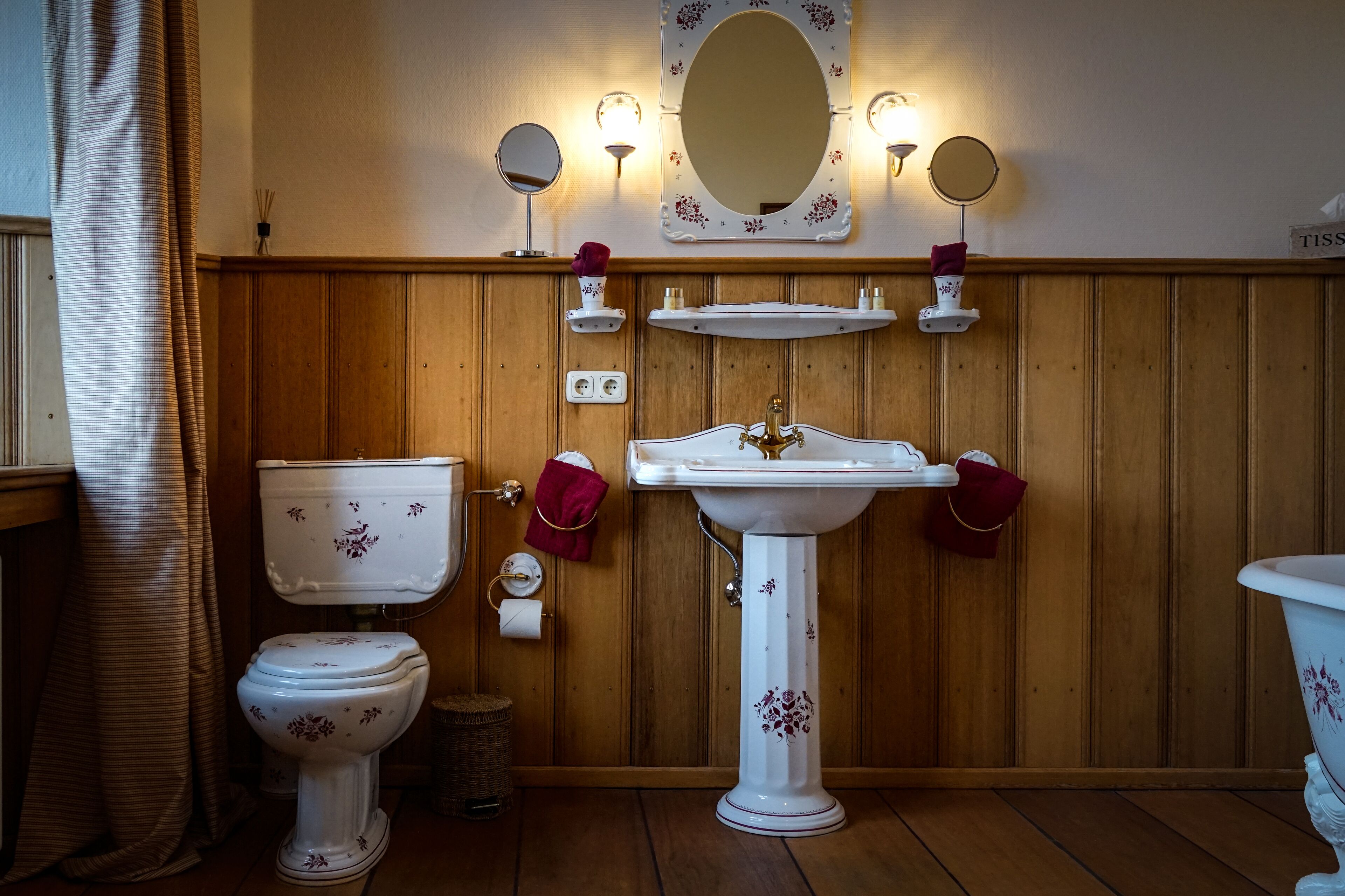 A bathroom with a wooden panel wall, a white pedestal sink, a toilet, and a mirror with wall sconces.