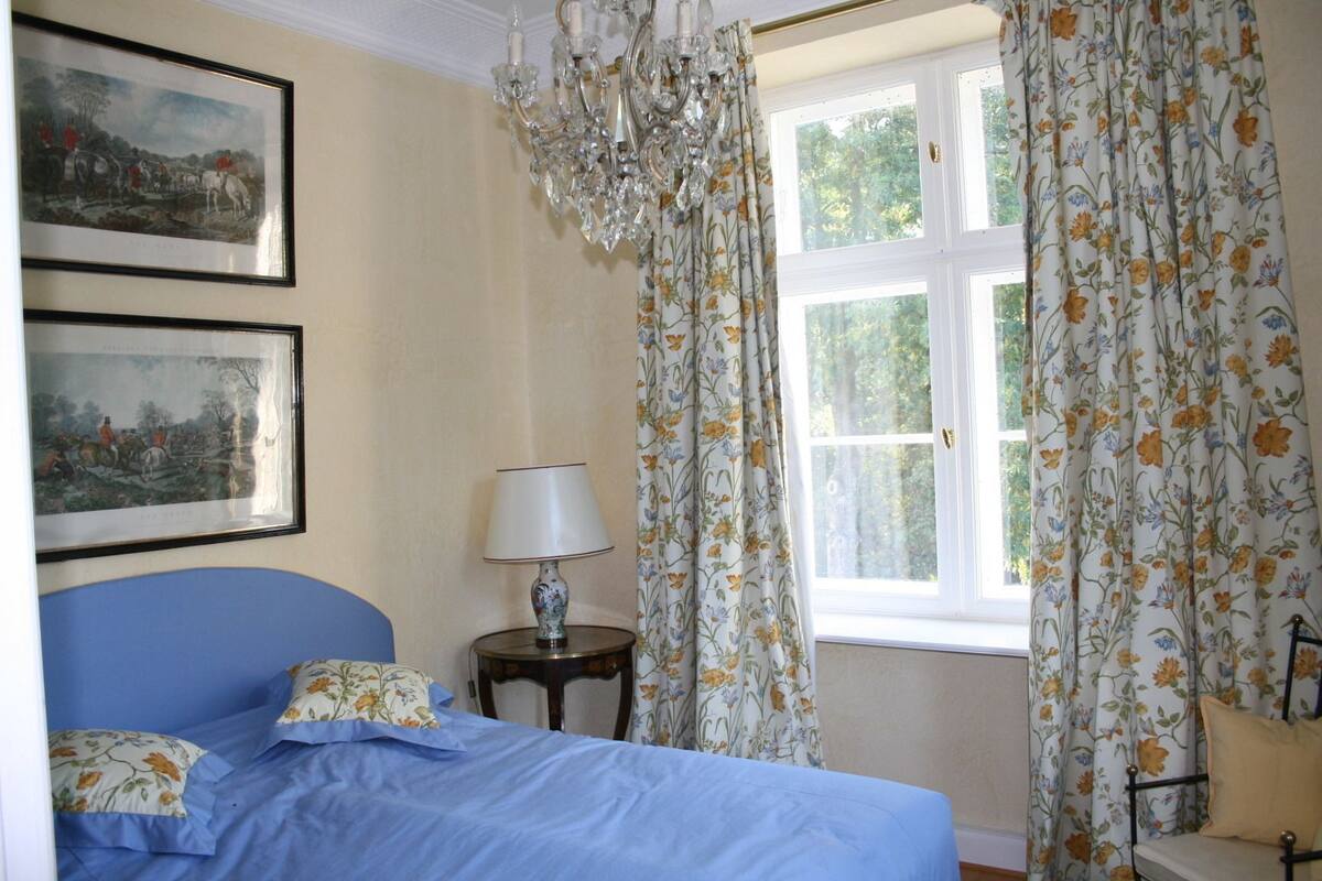 Un dormitorio con cama, mesita de noche, lámpara, cortinas con motivos florales y dos cuadros en la pared.