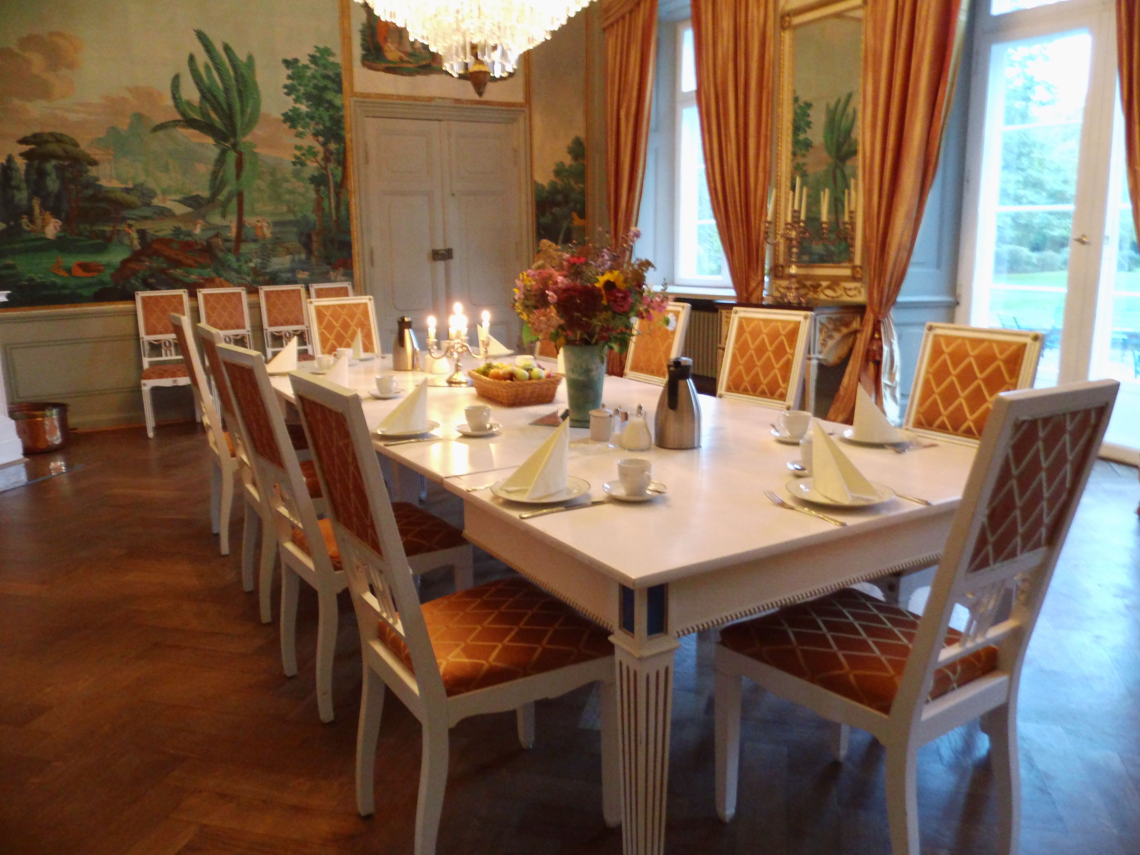Uma sala de jantar com uma mesa comprida posta para refeição, rodeada por cadeiras com assentos acolchoados.
