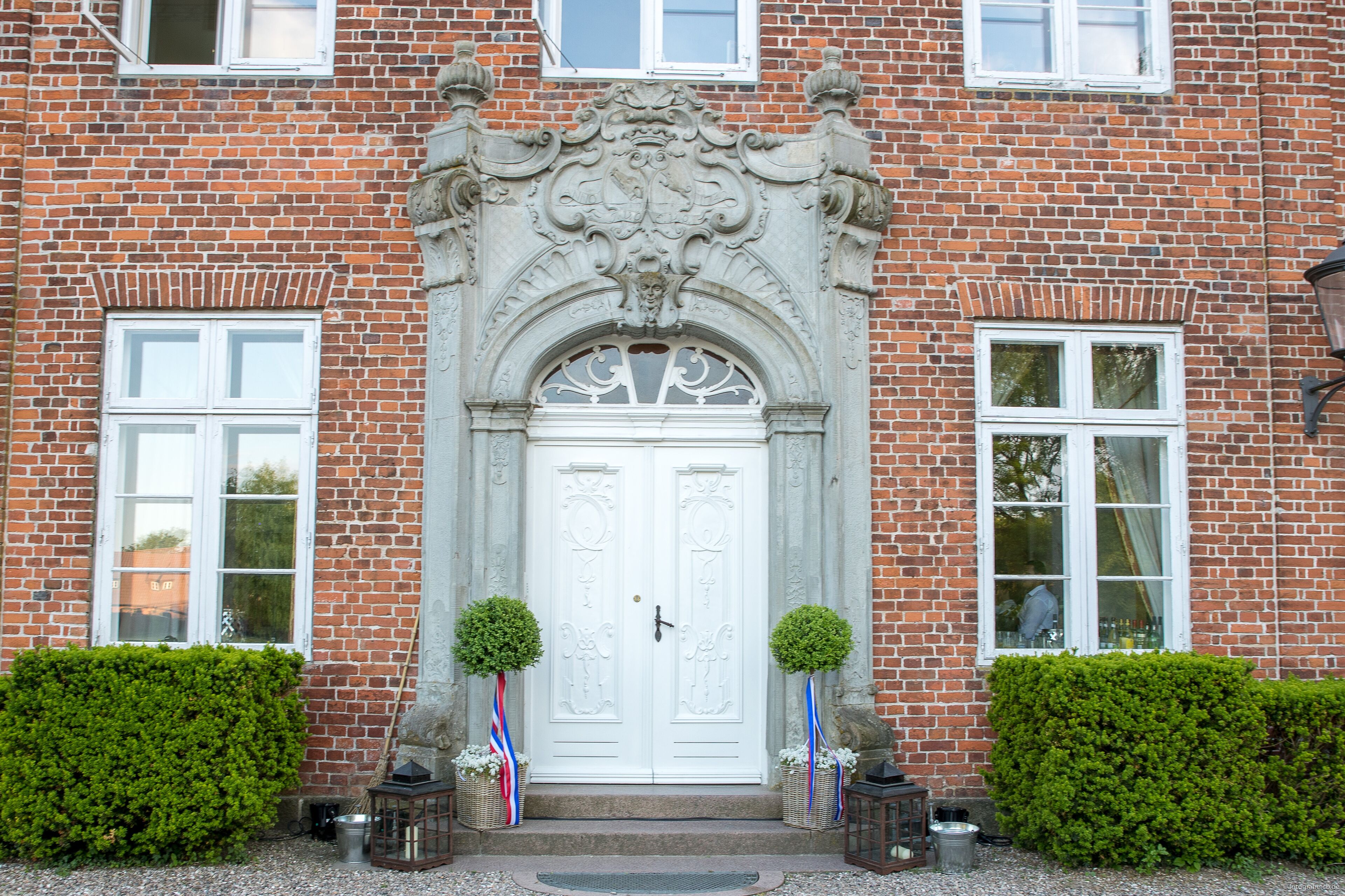 A brick building with a white ornate entrance door, two windows on either side, and a small step leading up to the door.